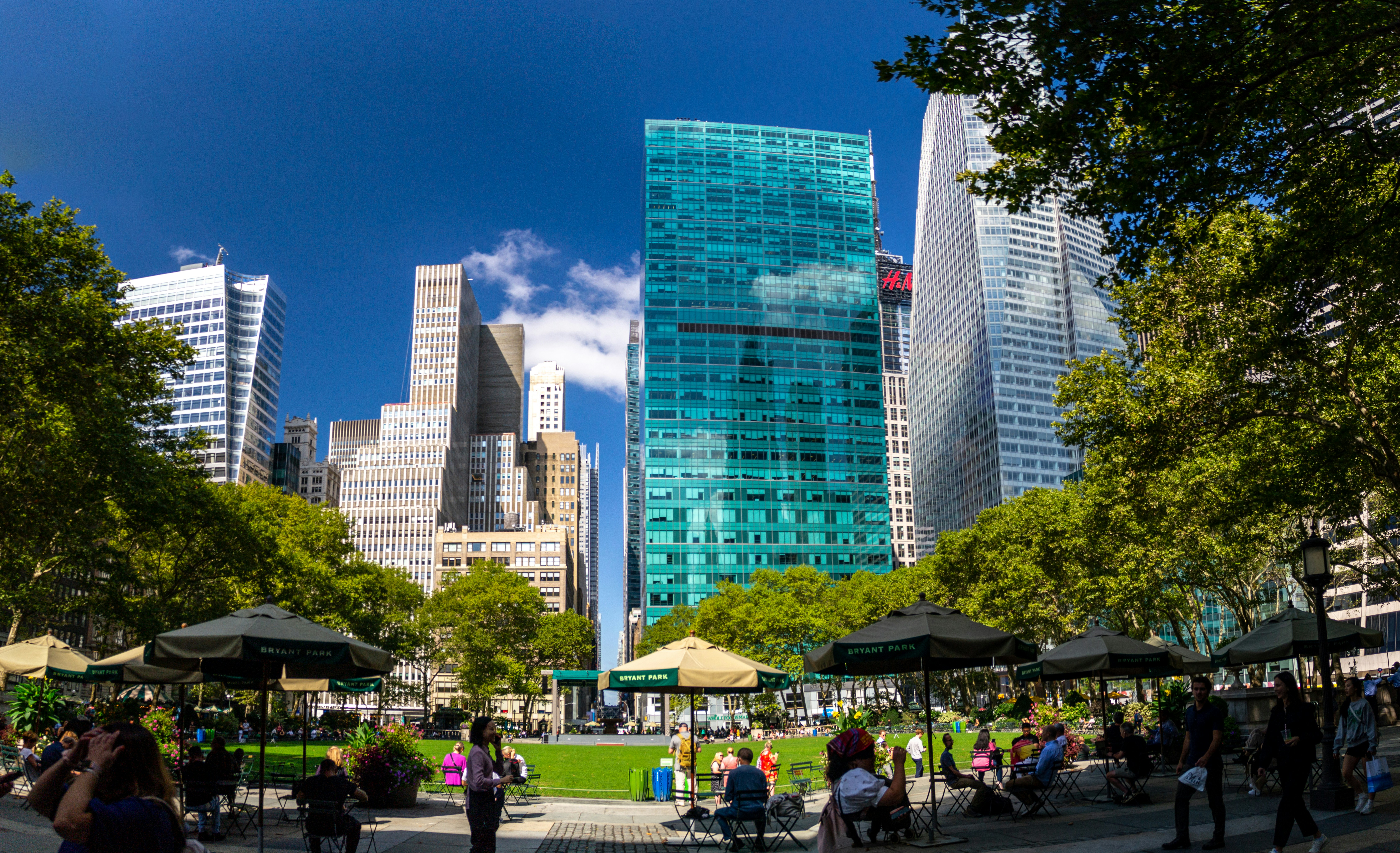 Imagen del parque de Bryant Park en el que se ven personas disfrutando de un buen clima, tomando café solos o acompañados. También se ven los edificios que rodean el parque, en el que destaca uno con los cristales azules.