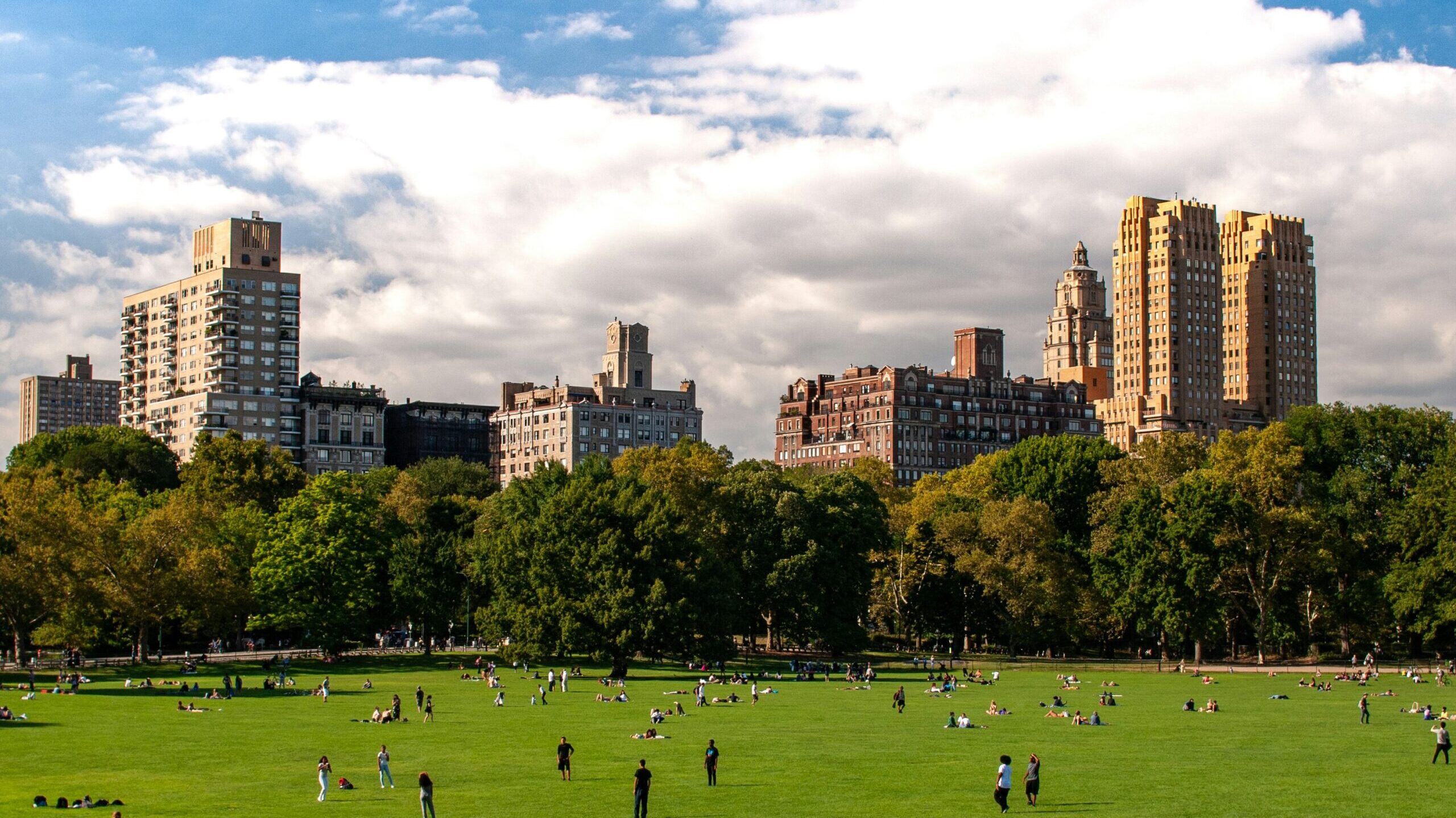 Imagen de Central Park de Nueva York con un tiempo muy soleado y se ve el césped con personas jugando a juegos, tumbadas o hablando con amigos. Se ven también los edificios que rodean el parque con unas nubes que hacen la imagen bonita.